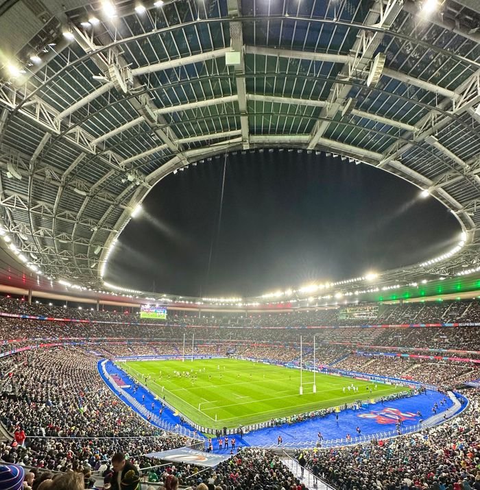 Nighttime panoramic view of a packed rugby match at Stade de France with dazzling lights and vibrant atmosphere.